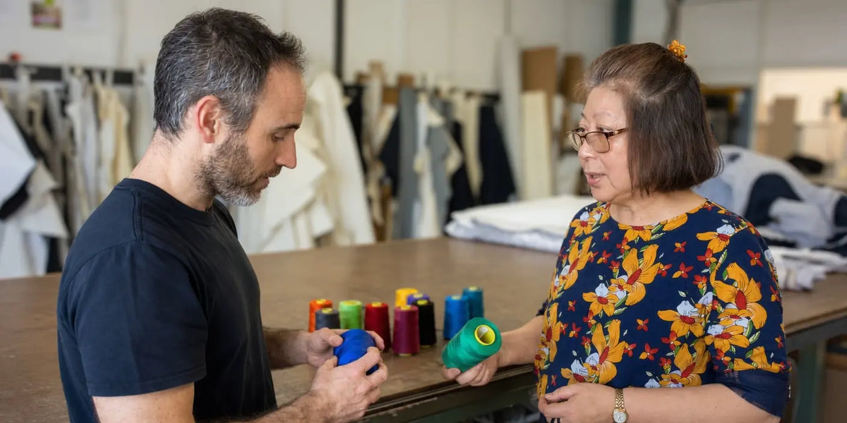 A man and a woman stand at a table in a workshop or studio, each holding a spool of thread. The man wears a dark navy T-shirt, and the woman wears a colorful floral-patterned blouse.