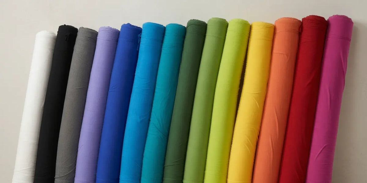A row of fabric rolls in a rainbow color sequence, including white, black, gray, purple, blue, teal, green, yellow, orange, red, and pink, displayed against a plain light background - these are representative of do+dare's variety of underwear colours.