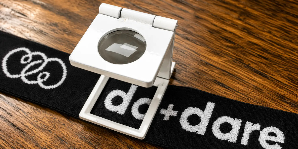 A white plastic magnifying glass holder attached to a black waistband with white text reading 'do+dare', placed on a wooden tabletop.