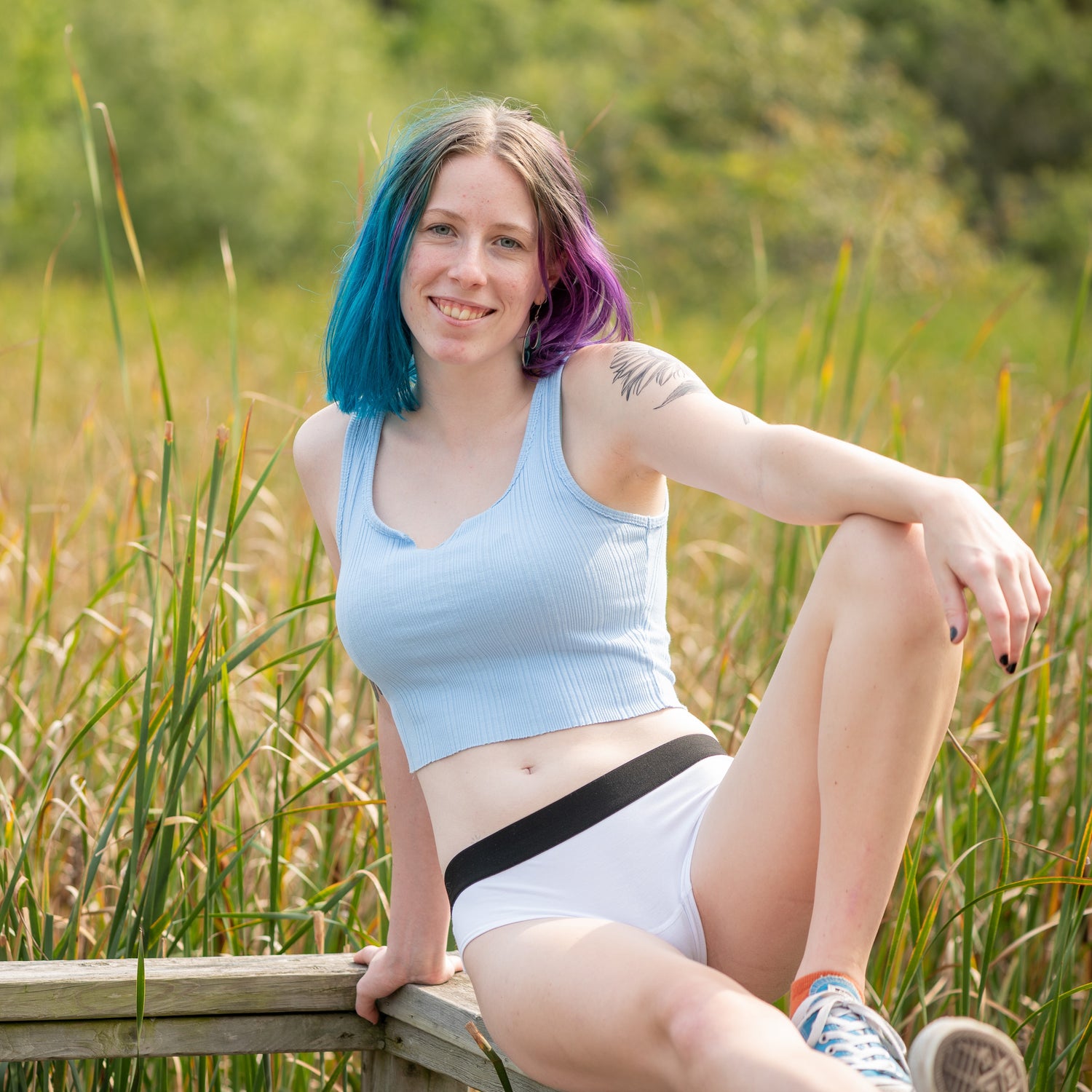 model with blue and purple hair sitting on a fence wearing light blue crop tank top and white do and dare cheeky underwear, smiling at camera